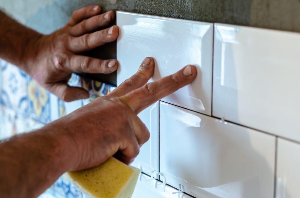 Hands of a tiler man gluing a white rectangular ceramic tile on a gray wall. Repair work indoors.