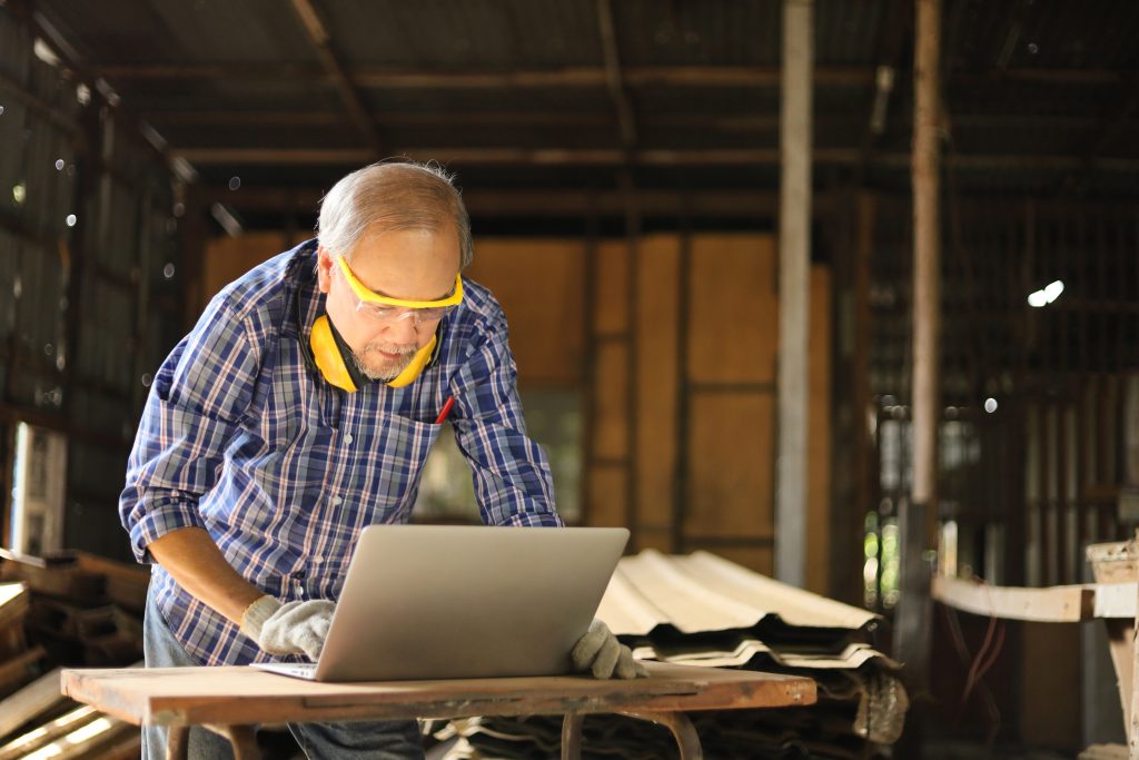 Senior Asian carpenter man is using computer laptop inside his home garage to get new modern inspiration and learning skill from online class and contacting international customers